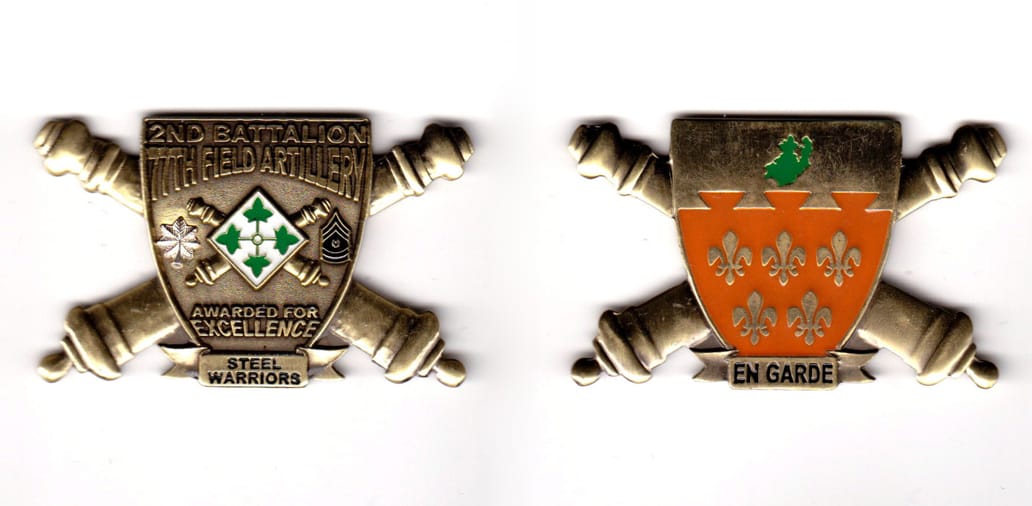 Shield shaped with crossed cannons extending, brass colored coin, 2-1/2 inches wide x 1-1/2 inches high.