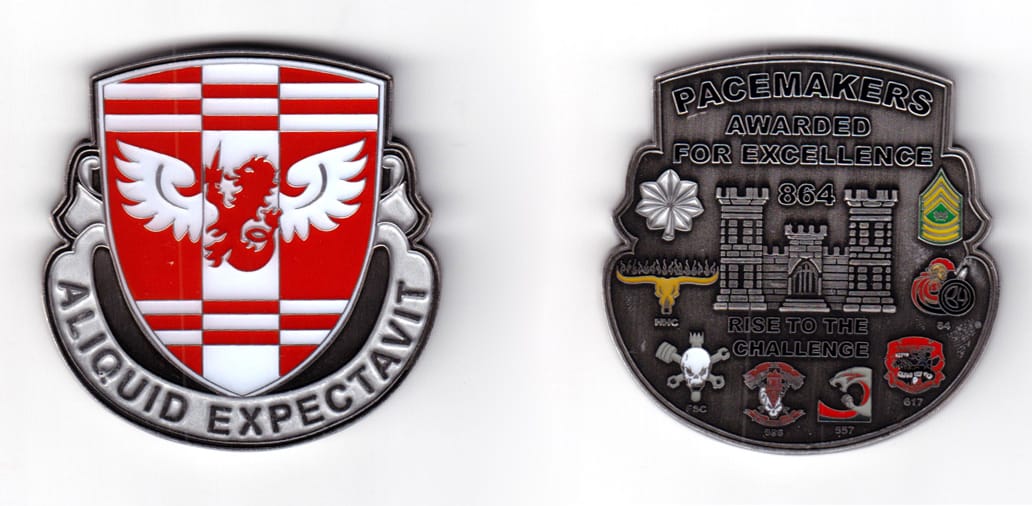 Shield shaped coin with a banner wrapped around the bottom 2/3 of the shield, aluminum in color, 2 15/64 inches high x 2...