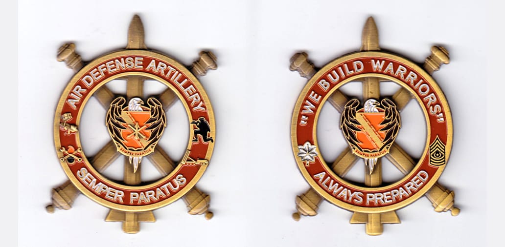 Round shaped coin backed by crossed artillery pieces and a vertical rocket, brass in color.